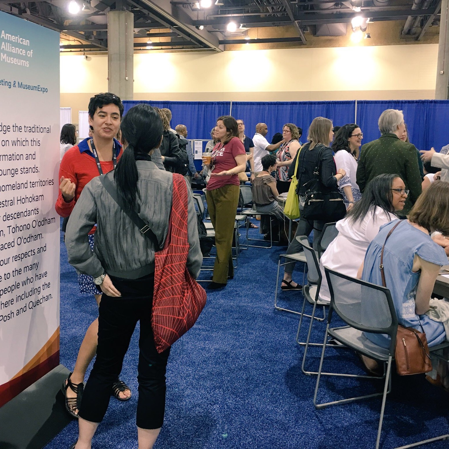 A crowd of a few dozen is gathered in various conversations, sitting and standing in small groups. The makeup of the crowd is diverse in age, gender, race, and ethnicity. A land acknowledgement standing banner for the American Alliance of Museums 2018 Annual Meeting is in the foreground.