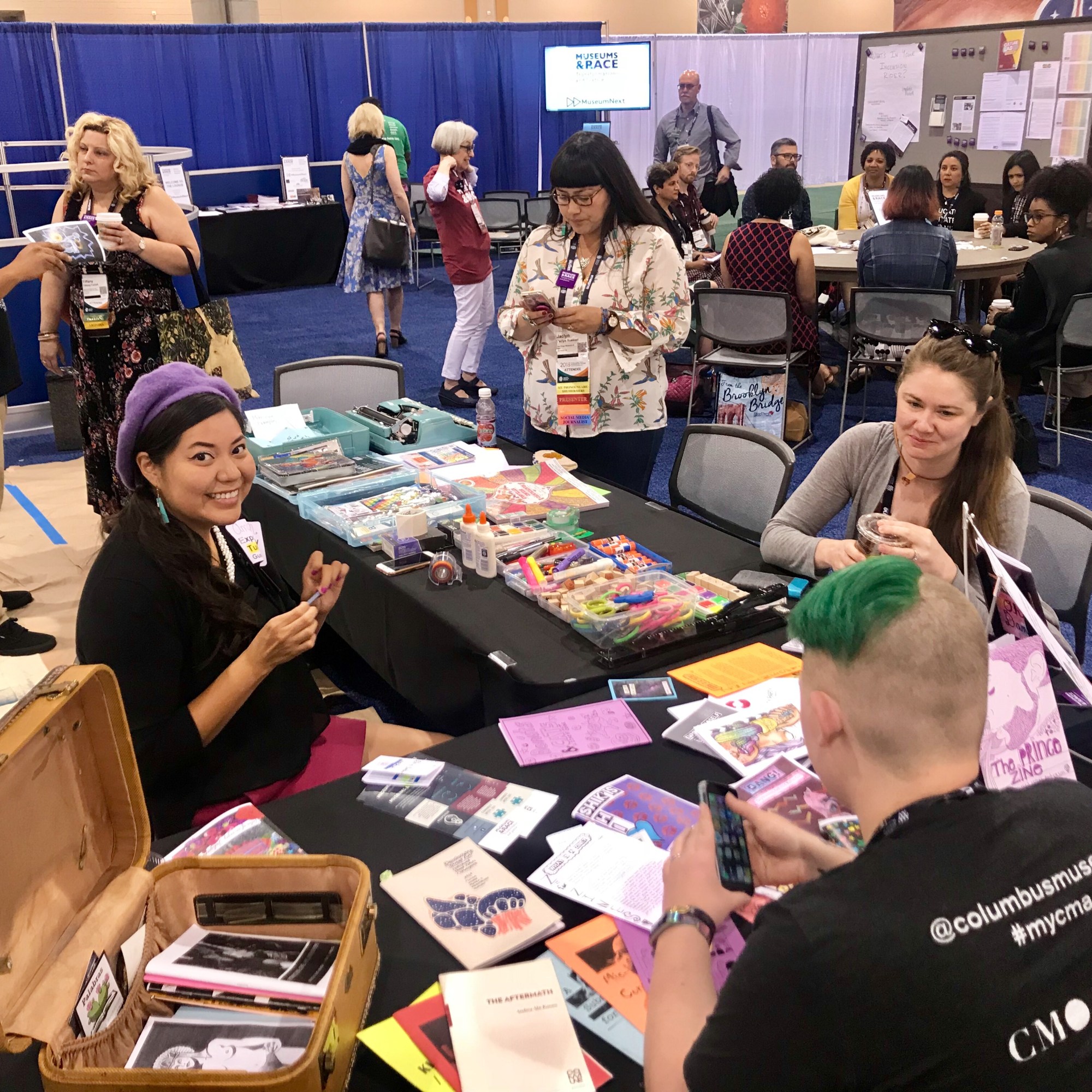 A racially diverse group of a few dozen people gather around tables in a conference hall. Four are around a table in the foreground covered in crafts, one of whom smiles at the camera. About a dozen participate in a round table discussion in the background. The others are scattered about standing in conversation or browser posted materials.