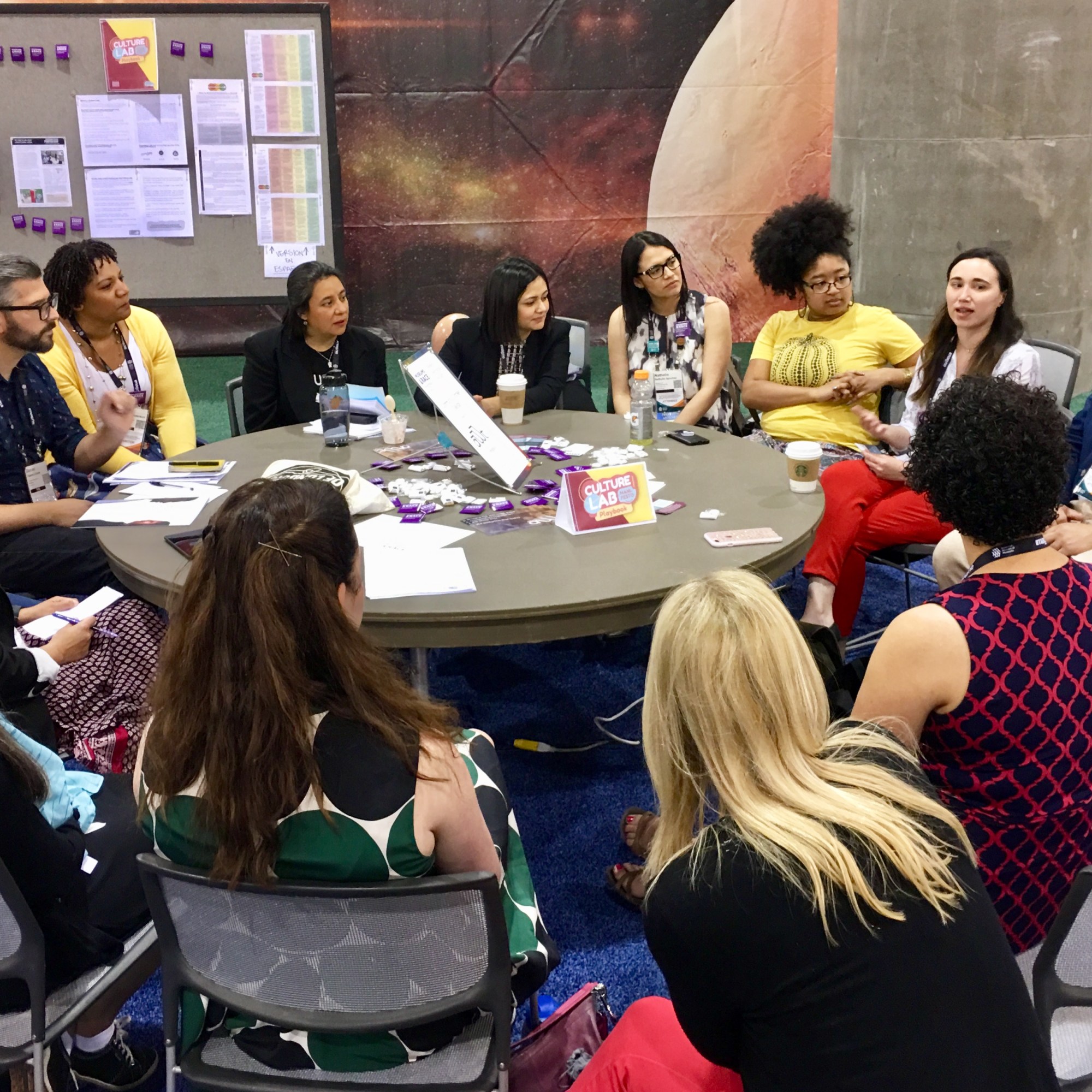 Seventeen people sit in close quarters around a round table. One person talks while the others listen intently. The group is diverse in apparent age, gender, race, and ethnicity. A few scattered materials are on the table and a sign that says "Culture Lab". A bulletin board is in the background covered with materials and buttons.