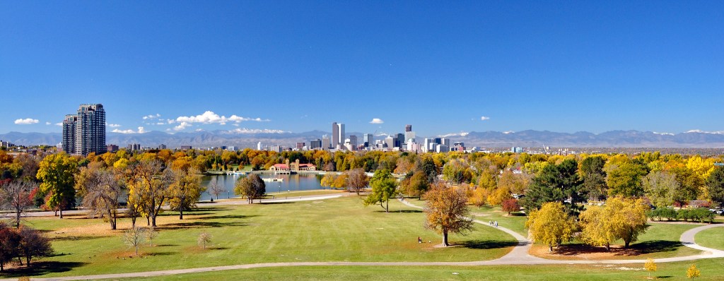 City skyline with a green expanse of park in the foreground, a lake and many leafy trees in the midground, a set of skyscrapers centered backed by a line of mountains with a bright blue sky arching over it all.