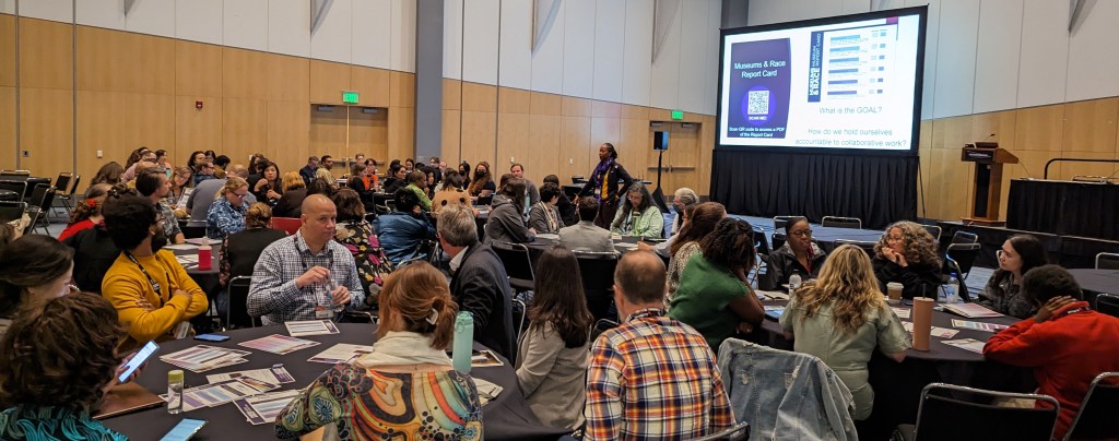 A set of conference roundtables packed with attendees. There are some papers visible on some of the tables and a slide is displayed in the background titled, "Museum & Race Report Card".