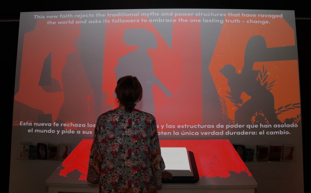 A person with shoulder length dark hair wearing a flower-print top looks at an exhibit panel with their back to the camera. The panel shows black silhouettes on a red background. The text on the panel says, "This new faith rejects the traditional yths and power structures that have ravaged the world and asks its followers to embrace the one lasting truth - change. A sentence in Spanish is at the bottom the panel, partially blocked by the observer.