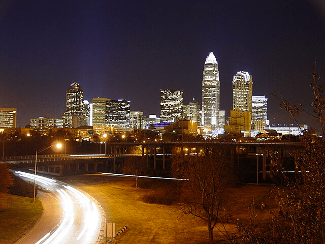 City skyline a night. Nearly a dozen skyscrapers are lit up shining brightly against a dark sky. A park dominates the foreground with highways crossing in between. A road curves from the left of the image to the bottom left corner with the streaks of headlights from an extended exposure tracking the paths of cars.