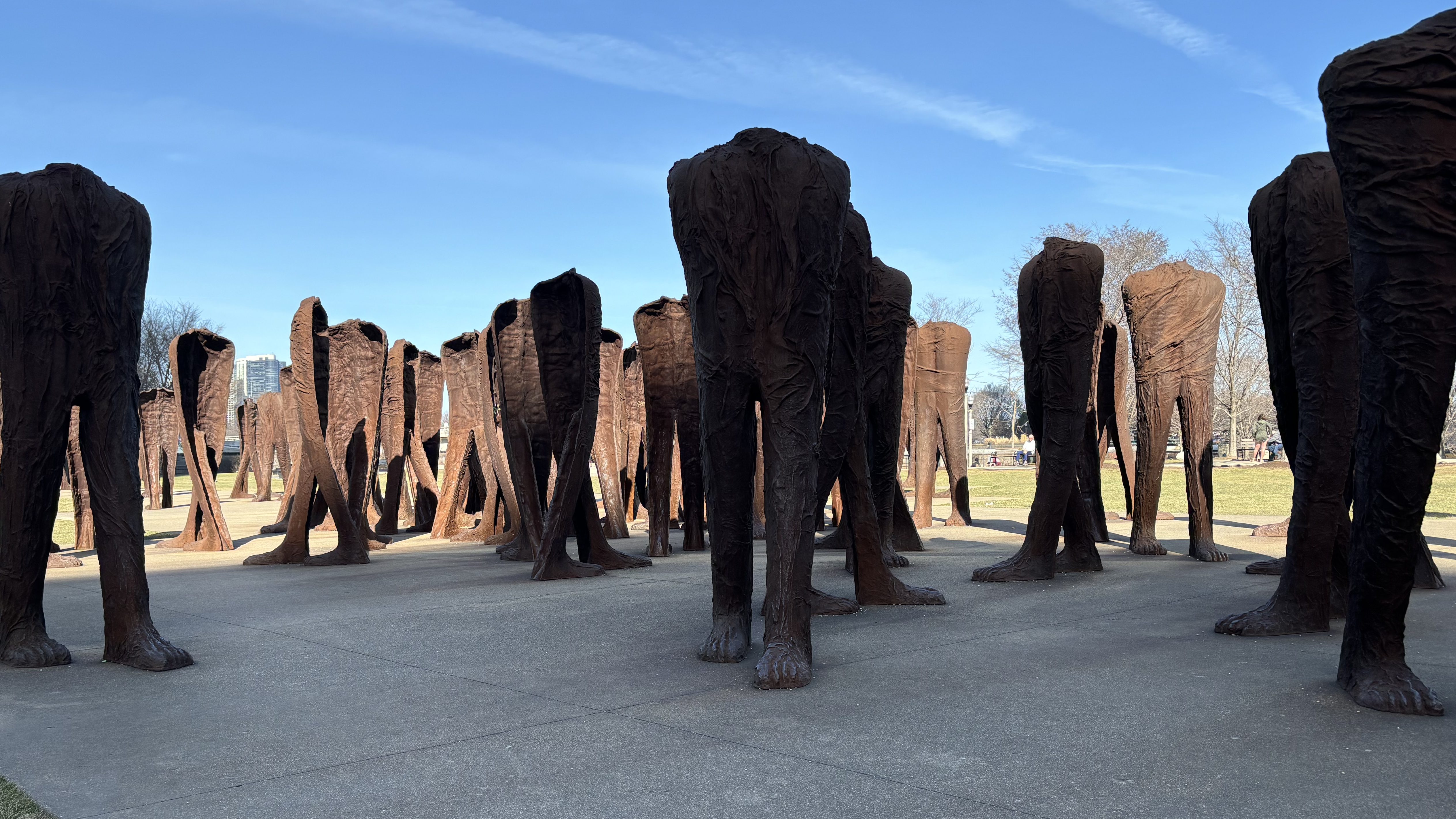 Multiple large headless and armless iron sculptures, appearing to be walking figures in multiple directions, installed in a park with blue sky, grass, and concrete 
