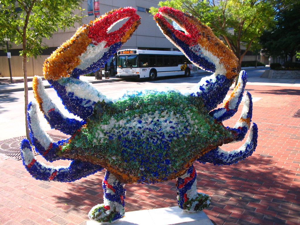 A crab stands upright on human-like feet, claws pointing towards the sky. The crab is covered in multi-colored crystals with blue and green dominating the body and it claws dominated by red and yellow. The crab sits on a brick patio with a bus visible in the background between the crab's claws.