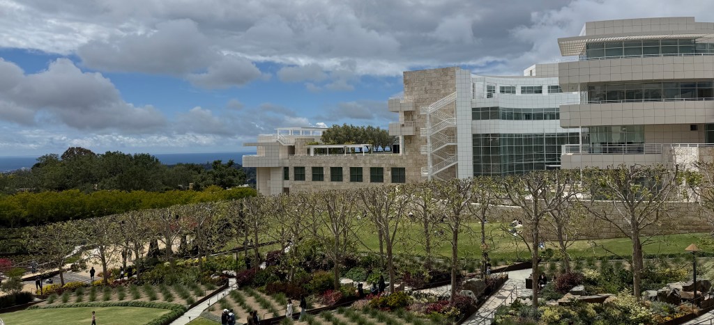 Landscape of the exterior of the Getty Museum Campus with the ocean in the distance. A row of starkly trimmed trees lines the foreground with barely visible buds of new growth.